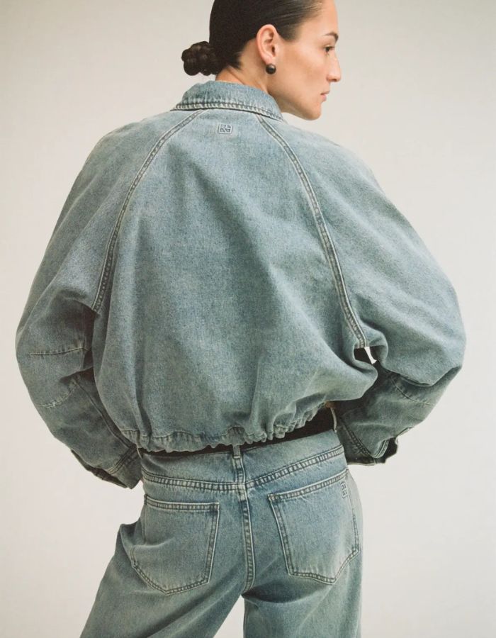Silhouette de dos avec le Blouson style bomber en jean pour femme de couleur bleu clair délavé Light Blue Denim, modèle Tiana de la marque Rouge Edit. Présente un col chemise évasé, une fermeture à glissière cachée sous des boutons-pression dorés mats Dull Gold et des brides de serrage ajustables.