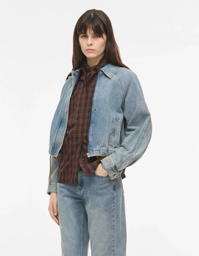 Silhouette avec le Blouson style bomber en jean pour femme de couleur bleu clair délavé Light Blue Denim, modèle Tiana de la marque Rouge Edit. Présente un col chemise évasé, une fermeture à glissière cachée sous des boutons-pression dorés mats Dull Gold et des brides de serrage ajustables.