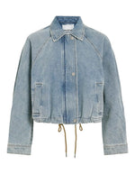 Blouson style bomber en jean pour femme de couleur bleu clair délavé Light Blue Denim, modèle Tiana de la marque Rouge Edit. Présente un col chemise évasé, une fermeture à glissière cachée sous des boutons-pression dorés mats Dull Gold et des brides de serrage ajustables.