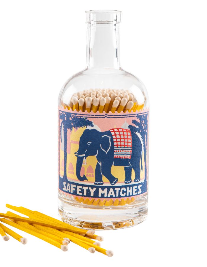 Grande bouteille en verre transparent avec bouchon en liège de la marque Archivist, servant de porte-allumettes. L'étiquette rétro montre un éléphant bleu sur fond de coucher de soleil rose. Le flacon contient de nombreuses allumettes en bois teinté jaune à tête blanche.
