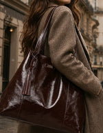 Silhouette avec le Grand sac cabas souple pour femme en cuir de vachette vernis bordeaux, modèle Odéon de la marque LOXWOOD. Présente deux anses de porté épaule, une patte de fermeture magnétique et un pompon amovible en cuir.