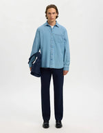 Homme portant une chemise en denim bleu clair Carlo, pantalon marine, et tenant un sac bleu.