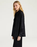 Profil du Chemisier long fluide noir pour femme au fini métallisé laminé brillant, modèle Bilancia Bis de la marque Momoni. Présente une coupe oversize avec de grands volants drapés, un col bateau asymétrique tombant sur l'épaule et des manches longues terminées par de hauts poignets à boutons recouverts.