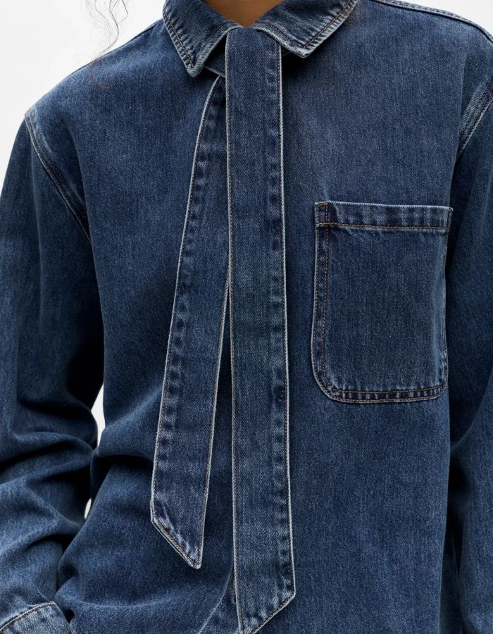 Detail de la Chemise en jean manches longues pour femme de couleur bleu moyen Medium Blue Denim, modèle Rosie de la marque Object. Confectionnée dans une toile de denim souple, elle présente un col chemise, une fermeture boutonnée, une poche plaquée sur la poitrine et un détail de nœud en tissu délicat.