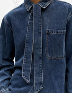 Detail de la Chemise en jean manches longues pour femme de couleur bleu moyen Medium Blue Denim, modèle Rosie de la marque Object. Confectionnée dans une toile de denim souple, elle présente un col chemise, une fermeture boutonnée, une poche plaquée sur la poitrine et un détail de nœud en tissu délicat.
