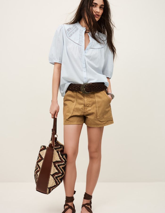 Chemise Nella Bleu Ciel en voile de coton biologique avec dentelle, manches courtes, portée avec un short et un sac.