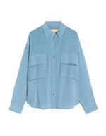 Chemisier oversize en pure soie de couleur bleu céruléen uni, modèle Pitanga de la marque Momoni. Présente une coupe ample et fluide avec un col bateau, une fermeture boutonnée originale sur le haut des épaules et des manches longues à poignets à la française.