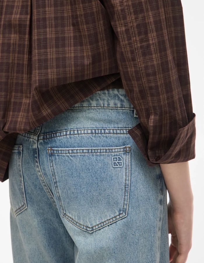 Bassin du Jean pour femme de couleur bleu clair délavé Light Blue Denim, modèle Tiana de la marque Rouge Edit. Présente une coupe droite ample contemporaine, une taille mi-haute avec des passants de ceinture et des finitions de boutons en métal doré mat Dull Gold.