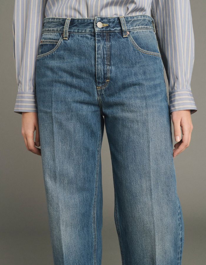 Ceinturedu Jean droit pour femme en denim bleu délavé avec plis marqués devant et dos, modèle Jerry de la marque SOEUR. Présente une coupe droite 5 poches, une fermeture boutonnée zippée et un patch en cuir au dos.