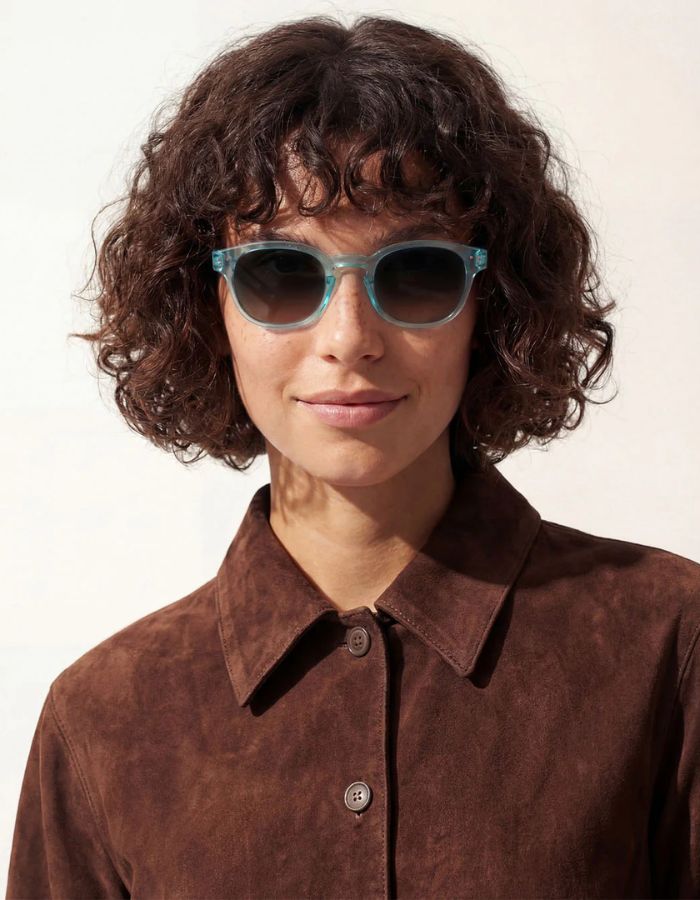 Femme avec les Lunettes de soleil unisexes IZIPIZI modèle #C de forme carrée vintage épaisse. La monture brillante est d'une couleur bleu turquoise translucide inspirée de la pierre turquoise, associée à des verres solaires bleus.