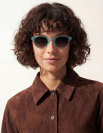 Femme avec les Lunettes de soleil unisexes IZIPIZI modèle #C de forme carrée vintage épaisse. La monture brillante est d'une couleur bleu turquoise translucide inspirée de la pierre turquoise, associée à des verres solaires bleus.