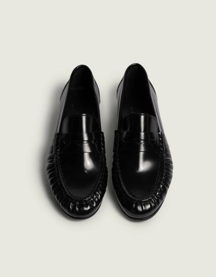 Dessus des Mocassins en cuir noir brillant pour homme et femme de type Penny Loafers, modèle Wrinkle de la marque POSA. Chaussures basses en cuir italien box poli avec une bride classique sur le coup de pied, un bout arrondi à empiècement plissé texturé et une semelle noire fine.