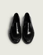 Dessus des Mocassins en cuir noir brillant pour homme et femme de type Penny Loafers, modèle Wrinkle de la marque POSA. Chaussures basses en cuir italien box poli avec une bride classique sur le coup de pied, un bout arrondi à empiècement plissé texturé et une semelle noire fine.