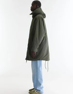 Profil de la Parka longue oversize pour homme couleur vert olive foncé avec capuche, fermeture zippée sous pression et poches en biais, modèle Jinx de la marque Bellerose.