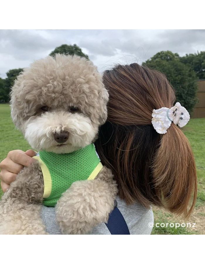 Mise en situation de la Petite pince à cheveux crabe de la marque Coucou Suzette en acétate de cellulose blanc nacré brillant. L'accessoire prend la forme d'un chien bichon frisé dodu portant un collier bleu turquoise avec des détails dorés.