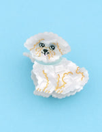 Petite pince à cheveux crabe de la marque Coucou Suzette en acétate de cellulose blanc nacré brillant. L'accessoire prend la forme d'un chien bichon frisé dodu portant un collier bleu turquoise avec des détails dorés.