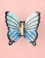 Pince à cheveux crabe de taille moyenne de la marque Coucou Suzette en acétate de cellulose brillant. L'accessoire prend la forme d'un papillon bleu azur et blanc nacré aux ailes déployées avec des contours noirs.