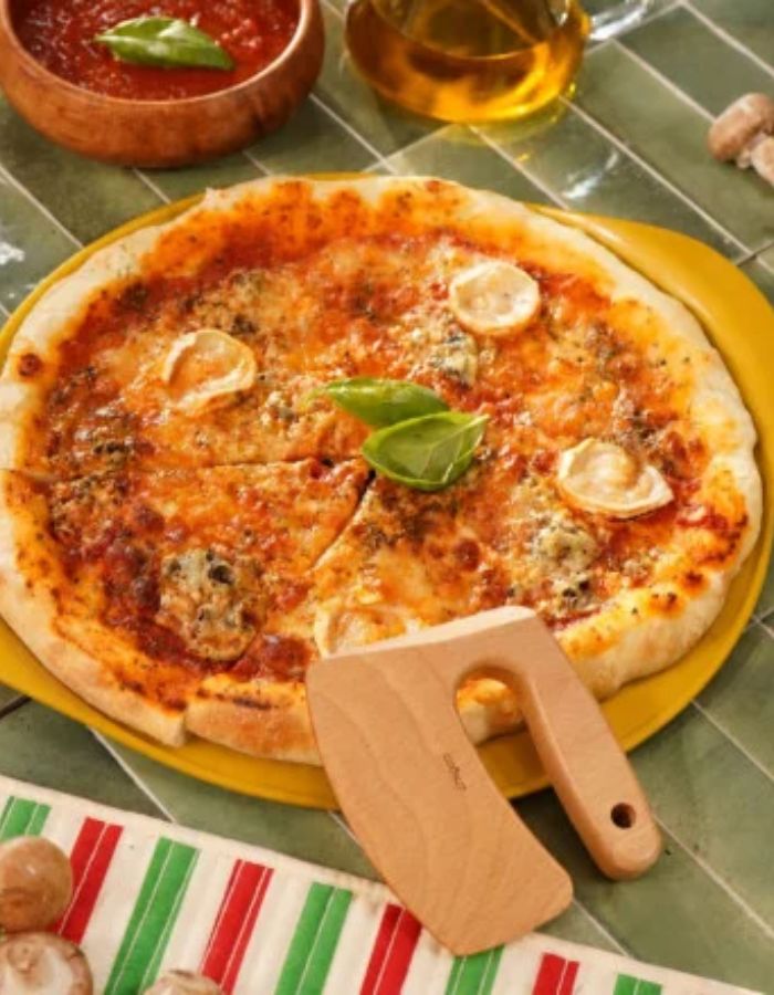 Le Formidable Plat à Pizza Cookut de 33 cm de diamètre, coloris jaune mangue. Plat de cuisson pour le four ultra-léger en métal recyclé avec revêtement antiadhérent, accompagné de son coupe-pizza en bois de hêtre.