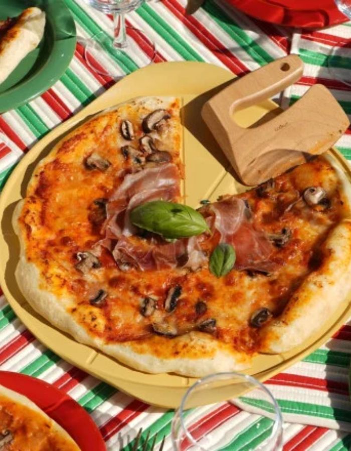 Le Formidable Plat à Pizza Cookut de 33 cm de diamètre, coloris jaune mangue. Plat de cuisson pour le four ultra-léger en métal recyclé avec revêtement antiadhérent, accompagné de son coupe-pizza en bois de hêtre.