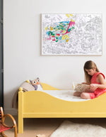 Fille lisant sur un lit jaune sous un grand poster à colorier Dinos OMY partiellement colorié.