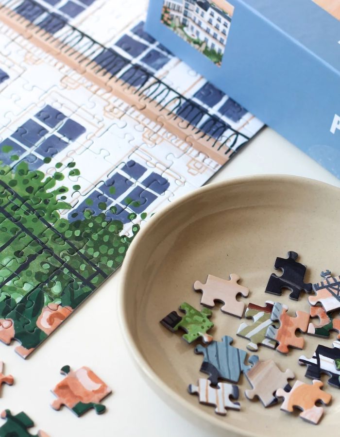 Détail de la Boîte de puzzle 1000 pièces Paris de la marque Piecely. L'illustration représente une table de petit-déjeuner sur un balcon avec croissants et café, offrant une vue romantique sur les toits de Paris et la Tour Eiffel au lever du soleil.