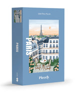 Boîte de puzzle 1000 pièces Paris de la marque Piecely. L'illustration représente une table de petit-déjeuner sur un balcon avec croissants et café, offrant une vue romantique sur les toits de Paris et la Tour Eiffel au lever du soleil.
