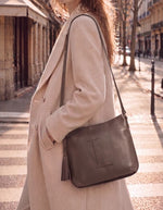 Femme portant le Sac besace pour femme en cuir grainé marron coffee avec logo L micro-perforé, pompon assorti et bandoulière réglable, modèle Celia L de la marque Loxwood.
