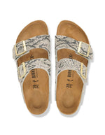 Dessus des Sandales pour pieds fins en largeur Narrow, modèle emblématique Arizona Rivet Logo de la marque Birkenstock. Confectionnées en cuir velours souple de couleur claire beige huître Snake Oyster, elles présentent deux brides horizontales ajustables avec boucles en métal, un rivet logo et la célèbre semelle anatomique marron en liège naturel