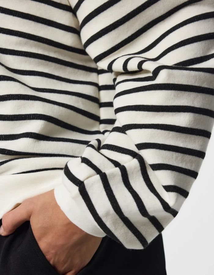Manche du Sweatshirt à col rond et manches longues pour femme de couleur écru blanc cassé Cloud Dancer, modèle Greta de la marque PIECES. Présente une coupe classique droite avec un petit cœur noir brodé sur le côté gauche de la poitrine.