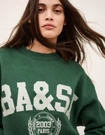 Femme portant un sweatshirt Fenja Emeraude Ba&sh vert foncé avec logo "BA&SH" blanc et motif campus.
