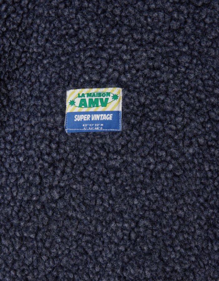 Logo de la Veste sans manches pour homme de couleur bleu nuit foncé Astre Chiné, modèle Hoktown de la marque American Vintage. Confectionnée dans une matière épaisse effet mouton retourné shearling, elle présente un col montant bas, une fermeture à boutons-pression, deux poches zippées et un patch logo sur la poitrine.