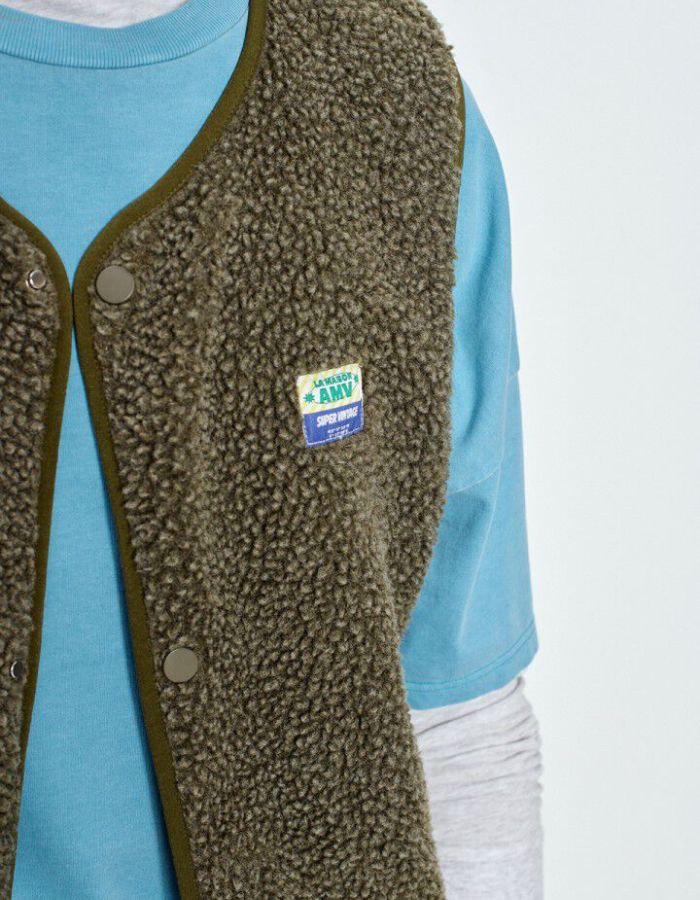 Col de la Veste sans manches pour homme de couleur vert kaki Varan Chiné, modèle Hoktown de la marque American Vintage. Confectionnée dans une matière épaisse effet mouton retourné shearling, elle présente un col montant bas, une fermeture à boutons-pression, deux poches zippées et un patch logo sur la poitrine.
