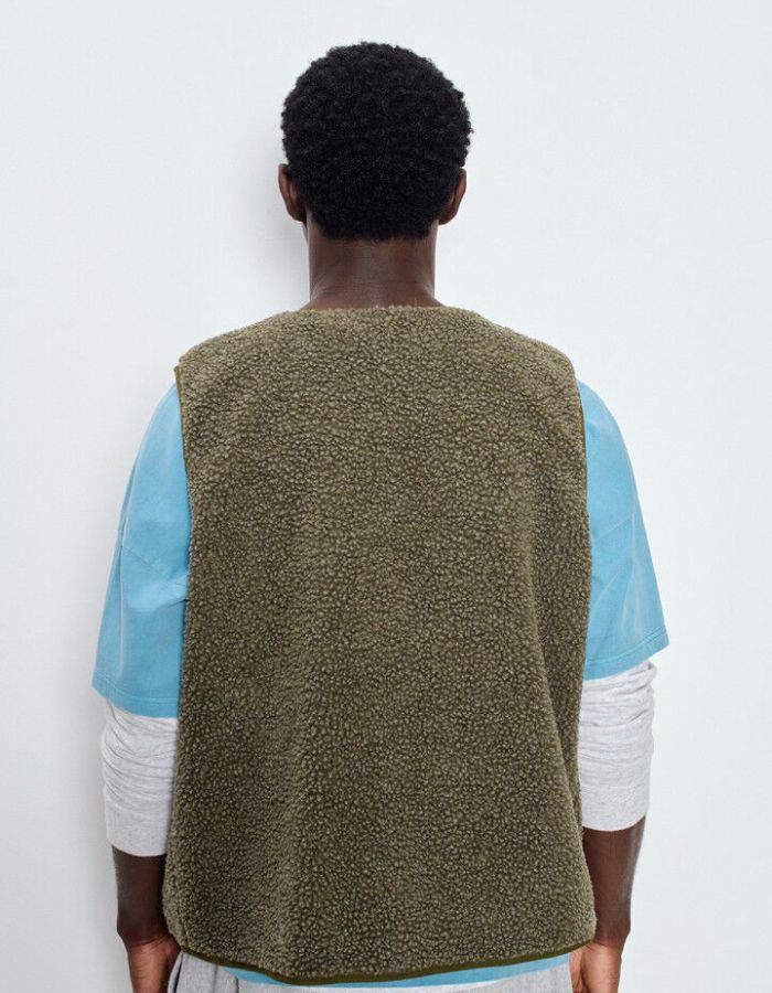Dos de la Veste sans manches pour homme de couleur vert kaki Varan Chiné, modèle Hoktown de la marque American Vintage. Confectionnée dans une matière épaisse effet mouton retourné shearling, elle présente un col montant bas, une fermeture à boutons-pression, deux poches zippées et un patch logo sur la poitrine.