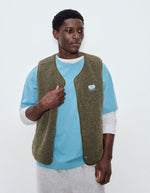 Vue de face de la Veste sans manches pour homme de couleur vert kaki Varan Chiné, modèle Hoktown de la marque American Vintage. Confectionnée dans une matière épaisse effet mouton retourné shearling, elle présente un col montant bas, une fermeture à boutons-pression, deux poches zippées et un patch logo sur la poitrine.