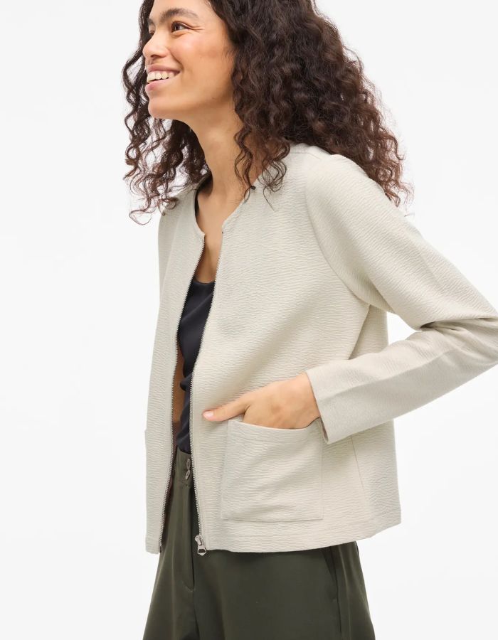 Femme souriante portant une veste beige zippée Garden Vila en maille côtelée, coupe courte, col rond et poches avant.